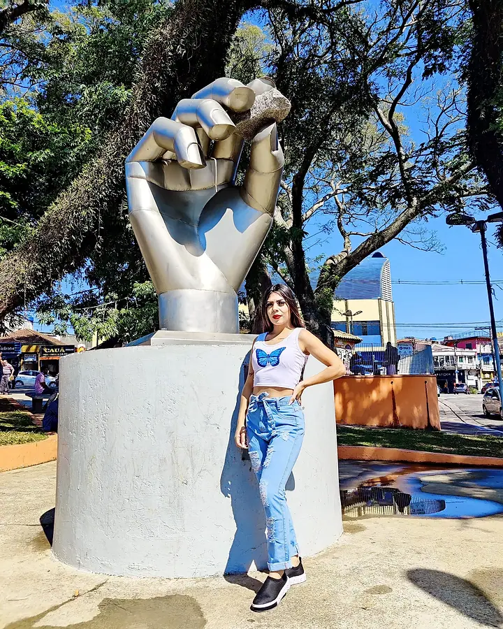 Modelo de cropped branco e calça jeans ao lado de uma escultura de mão numa praça